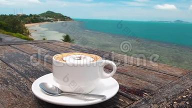 白色杯子里的风味咖啡，放在盘子里，用勺子放在木制的乡村桌子上，海的背景和棕榈树摇曳着